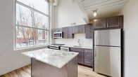 Located in the heart of downtown Columbus, this two-floor urban loft at 60 E Spring St blends industrial character with modern upgrades. The main level showcases soaring ceilings with exposed ductwork and large floor-to-ceiling windows that flood the space with natural light and offer views of the surrounding cityscape. Light hardwood flooring runs throughout the open-concept living and kitchen areas, creating a warm, cohesive aesthetic. The kitchen is a standout feature, equipped with dark shaker-style cabinetry, stainless steel appliances, a marble-topped center island, subway tile backsplash, and a deep utility sink. A slatted wood ceiling detail adds architectural interest at the transition to the hallway and upper-level staircase. The bathroom is thoughtfully finished with a dark-framed vanity mirror, granite countertop, industrial-style sconce lighting, and a tub-shower combination. An in-unit stacked washer and dryer provides added convenience. The lower level offers carpeted flex spaces with exposed concrete ceilings, structural columns, and visible HVAC infrastructure — ideal for use as additional living, office, or storage areas. Together, the two floors offer a dynamic, loft-style living experience in a prime downtown Columbus location.