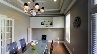 This single-floor space exudes a polished, functional aesthetic with thoughtful architectural details throughout. The main room showcases a tray ceiling with a recessed border, adding depth and a sense of elevated style to the interior. A statement chandelier with classic black shades and ornate metalwork serves as a focal point overhead, casting a warm, sophisticated ambiance across the room.

The walls are finished in a neutral warm gray tone, complementing the rich hardwood flooring that runs throughout the space. Built-in white cabinetry lines one wall, combining upper enclosed cabinets with open display shelving — offering both storage and the opportunity to showcase decorative items. A dedicated lower counter section provides additional workspace or display surface below the shelving unit.

Natural light filters in through a window fitted with white horizontal blinds, brightening the space while allowing for privacy control. A glass-top dining or conference table with sleek black chairs occupies the center of the room, making it well-suited for meetings, dining, or collaborative work. The combination of classic architectural features, built-in storage, and contemporary furnishings gives this space a versatile and refined character.