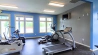Located at 3995 Cracker Cover Ln in Canal Winchester, Ohio, this single-floor fitness center is designed to support an active lifestyle with a clean, functional layout. The space features a bold color scheme, with a vibrant blue accent wall complemented by neutral gray walls, creating an energizing atmosphere conducive to exercise. Large windows along the front wall allow ample natural light to flood the room while offering views of the surrounding greenery, enhancing the overall workout experience. The facility is equipped with a selection of cardio machines, including treadmills and elliptical trainers, all positioned to take advantage of the natural light and outdoor views. A wall-mounted flat-screen television provides entertainment during workouts. Durable, dark rubberized flooring runs throughout the space, offering both safety and comfort underfoot. Overhead fluorescent lighting ensures the room remains bright and well-lit at all times. The space is thoughtfully arranged to allow for comfortable movement between equipment, making it accessible and practical for users of all fitness levels.