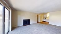 This single-floor residence offers a straightforward and functional layout designed for comfortable everyday living. The main living area is carpeted and centers around a classic fireplace framed in dark surround, providing a focal point and a cozy atmosphere for relaxing or entertaining. The neutral, light-toned walls and textured ceiling contribute to a clean, understated aesthetic throughout the space. Transitioning seamlessly from the living area, the kitchen features white cabinetry that extends to the upper walls, offering ample storage. Appliances including a microwave and range are integrated into the kitchen layout, and the flooring shifts to a warm hardwood tone, providing a pleasant contrast to the carpeted living space. A doorway visible from the main area leads to additional rooms or storage, suggesting a practical flow throughout the unit. The overall ambiance is calm and neutral, presenting a versatile canvas well-suited for personalization. The open sightlines between the living and kitchen areas enhance the sense of space, making the interior feel connected and easy to navigate.