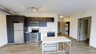 This single-floor unit offers a thoughtfully designed open-concept layout that maximizes both functionality and style. The kitchen serves as the centerpiece of the space, featuring dark espresso-toned cabinetry with clean-lined hardware, contrasting light countertops, and a classic white subway tile backsplash that adds a timeless touch. A full suite of stainless steel appliances — including a refrigerator, electric range, and over-the-range microwave — provides a sleek, cohesive look. A movable kitchen island with a white surface offers additional prep space and flexibility. The sink is positioned beneath upper cabinetry, maximizing storage throughout. Light-toned laminate flooring runs throughout the unit, creating a warm and cohesive aesthetic that complements the modern design palette. The open layout flows naturally from the kitchen into the living area, allowing for versatile furniture arrangements. A dedicated in-unit laundry area is conveniently tucked into its own alcove, housing a stacked washer and dryer. Recessed and track lighting provides bright, even illumination throughout. This well-appointed unit combines practical design with contemporary finishes, making it a comfortable and functional living space.