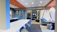 This well-appointed single-floor space showcases a thoughtfully designed interior that blends modern aesthetics with functional comfort. The open-concept layout connects a dining and lounge area seamlessly, creating an inviting environment ideal for socializing and relaxation.

The dining area features a sleek white countertop island with contemporary gray seating, providing a clean and practical gathering spot. Bold design choices are evident throughout, with a striking orange ceiling accent and rich blue accent walls that add depth and character to the space. Abstract artwork adorns the walls, contributing to the curated, modern atmosphere.

Beyond the dining area, the lounge zone is anchored by a stylish fireplace framed in dark tile, flanked by built-in bookshelves that offer both storage and display space. A wall-mounted television sits above the fireplace, making this area a natural hub for entertainment. Comfortable dark upholstered seating and a window nook with teal accent cushions provide additional lounging options.

Recessed lighting throughout ensures the space is well-lit, while light wood-toned flooring and neutral gray tones balance the vibrant accent colors. The overall ambiance is modern, dynamic, and welcoming — a versatile communal space designed with both style and practicality in mind.