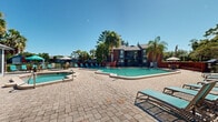 Located at 675 Jamestown Blvd in Altamonte Springs, Florida, this two-story residential community offers a well-appointed outdoor amenity area designed for relaxed Florida living. The centerpiece of the outdoor space is a generously sized swimming pool with crystal-clear turquoise water, complemented by a separate octagonal spa positioned adjacent to the main pool. The pool deck is finished with attractive paver stonework and is outfitted with aqua-blue lounge chairs and chaise lounges, providing ample space for sunbathing and outdoor relaxation. Multiple patio umbrellas offer shaded seating areas throughout the deck. A dedicated clubhouse or amenity building sits alongside the pool area, featuring large windows and a pitched roof, serving as a functional hub for residents. The community is framed by mature tropical landscaping, including tall palm trees and dense native foliage, lending a lush, resort-like ambiance. The two-story residential buildings feature classic brick exteriors with balconies and stairwell access, visible in the background and integrated naturally into the landscaped setting. Under the consistently sunny Central Florida skies, this outdoor area delivers a vibrant and inviting atmosphere throughout the year.