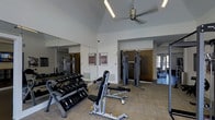 Located at 2234 South Trenton Way in Denver, Colorado, this fitness center offers a well-equipped, single-floor workout facility designed to support a comprehensive exercise routine. The space features soaring vaulted ceilings that lend an open, airy feel, complemented by a ceiling fan that promotes air circulation throughout workouts. Large floor-to-ceiling mirrors line one wall, visually expanding the space and allowing for proper form checks during exercise. Natural light filters in through glass doors, creating a bright and energizing atmosphere. The facility is outfitted with a full dumbbell rack offering a range of weights, an adjustable weight bench, and cable resistance machines suited for a variety of strength training exercises. Additional cable towers are visible in the adjoining area, further extending the range of available equipment. The flooring features durable, textured carpeting appropriate for a high-traffic fitness environment. Informational signage is posted on the walls, providing guidance and facility rules for users. The layout is thoughtfully organized to allow for comfortable movement between equipment stations, making this fitness center a practical and functional amenity for residents and users of the surrounding community.