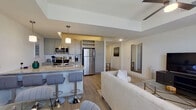 This well-appointed single-floor residence showcases a thoughtfully designed open-concept layout that seamlessly connects the kitchen, dining, and living areas. The kitchen is a functional centerpiece, equipped with stainless steel appliances including a full-size refrigerator, electric range, and microwave. White cabinetry, granite countertops, and a spacious island with bar seating and pendant lighting above provide both style and practicality. A built-in wine rack adds a distinctive touch. Light wood-style flooring flows throughout the main living areas, contributing to a clean, cohesive aesthetic.

The living room is generously proportioned, featuring a striking arched window that draws in abundant natural light. A ceiling fan, glass coffee table, and comfortable sectional sofa furnish the space, while an adjacent dining area accommodates a round glass table with seating. The bedroom offers plush carpeting, a tufted upholstered headboard, and a dedicated walk-in closet with wire shelving. A well-lit hallway with a circular mirror connects the rooms efficiently. Throughout, the neutral color palette, recessed lighting, and modern fixtures create a calm, contemporary ambiance.