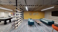 This community room showcases a compelling fusion of industrial and contemporary design across a single, open floor. The space is anchored by an exposed ceiling that features a striking grid of horizontal wood slats suspended from black steel beams, complemented by linear LED lighting and visible ductwork and piping that reinforce the industrial aesthetic. Original cast-iron structural columns add historic character throughout.

The main lounge area is divided into distinct seating zones by a dramatic freestanding slatted wood partition, which creates visual separation without sacrificing openness. Teal and aqua-toned sofas arranged around a low wood coffee table invite relaxed gathering, while a bold mustard-yellow accent wall adorned with curated artwork energizes the space. A high-top bar table with orange stools provides an additional casual seating option.

The kitchenette is well-appointed with granite countertops, white cabinetry, a built-in wall oven, dishwasher, microwave, and a decorative tile backsplash — designed to support events and gatherings. Pendant cage lights add warmth to this functional area.

Underfoot, light gray wood-look flooring with teal accent streaks ties the design palette together cohesively. The overall ambiance is vibrant, modern, and welcoming, making this community room well-suited for social events, meetings, and everyday gatherings.