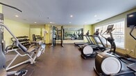 Located at 215 Fairmount Avenue in Lynn, Massachusetts, this well-equipped fitness center offers a comprehensive workout facility across a single floor. The space is thoughtfully designed with light yellow-green walls, recessed ceiling lighting, and durable rubber flooring suited for high-traffic exercise use. Natural light filters in through multiple windows along the perimeter, creating a bright and energizing atmosphere. Large wall-mounted mirrors span a significant portion of the room, visually expanding the space and allowing users to monitor their form during workouts. The cardio area is outfitted with several treadmills and elliptical machines, each equipped with individual digital displays. A Precor cable cross-trainer serves as a centerpiece for strength training, complemented by a dedicated free weight section featuring a multi-tiered dumbbell rack. Additional strength and resistance machines are positioned throughout the room, alongside a collection of exercise and stability balls stored on a wall-mounted rack. A flat-screen television is mounted on the wall for entertainment during workouts. The facility also includes a storage closet and a glass-paneled entry door with an illuminated exit sign, ensuring safe and convenient access. Overall, the fitness center provides a functional, well-organized environment suited for a full range of exercise routines.