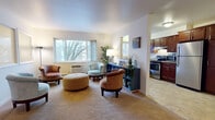 This deluxe 2-bedroom, 1-bathroom residence is laid out entirely on a single floor, offering a practical and comfortable living arrangement. The spacious living room serves as the heart of the home, featuring neutral carpet flooring, bright white walls, and a large picture window that frames outdoor tree views and fills the room with natural light. The open arrangement accommodates a generous seating area, complemented by a wall-mounted air conditioning unit and baseboard heating.

Adjacent to the living room, the kitchen is outfitted with rich dark wood cabinetry, stainless steel appliances including a refrigerator and range, and tile flooring that adds a clean, functional contrast to the carpeted living areas. The kitchen connects seamlessly to the main living space, making the layout feel open and easy to navigate.

Both bedrooms are carpeted and well-lit by natural light through sizeable windows. Each room offers ample floor space and includes closet storage. The rooms maintain a calm, neutral aesthetic with clean white walls.

The bathroom features a granite-topped vanity with dark wood cabinetry, white subway tile surround, a tub-shower combination, and tile flooring, creating a polished and cohesive finish throughout.