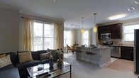 Located at 243 Steele, this single-floor residence offers an open-concept layout that integrates the living, dining, and kitchen areas into one cohesive and functional space. Large windows dressed with sheer gold-toned curtains flood the interior with abundant natural light, creating a warm and inviting ambiance throughout.

The living area features neutral-toned carpeting and is anchored by a charcoal sofa and a sleek glass-top coffee table, contributing to a contemporary yet comfortable aesthetic. Crown molding adds a refined architectural detail along the ceiling line.

The kitchen is a standout feature, showcasing dark espresso cabinetry, stainless steel appliances including a microwave and range, and a light-toned granite or stone countertop island. Pendant lighting hangs above the island, which provides additional seating with bar-height chairs, making it ideal for casual dining or entertaining.

Adjacent to the kitchen, a dedicated dining area accommodates a small table and chairs, bathed in natural light from the surrounding windows. Track lighting overhead ensures the space is well-lit at all times. The overall design emphasizes clean lines, neutral tones, and an airy, modern feel.