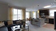 Located at 243 Steele, this single-floor residence offers an open-concept layout that integrates the living, dining, and kitchen areas into one cohesive and functional space. Large windows dressed with sheer gold-toned curtains flood the interior with abundant natural light, creating a warm and inviting ambiance throughout.

The living area features neutral-toned carpeting and is anchored by a charcoal sofa and a sleek glass-top coffee table, contributing to a contemporary yet comfortable aesthetic. Crown molding adds a refined architectural detail along the ceiling line.

The kitchen is a standout feature, showcasing dark espresso cabinetry, stainless steel appliances including a microwave and range, and a light-toned granite or stone countertop island. Pendant lighting hangs above the island, which provides additional seating with bar-height chairs, making it ideal for casual dining or entertaining.

Adjacent to the kitchen, a dedicated dining area accommodates a small table and chairs, bathed in natural light from the surrounding windows. Track lighting overhead ensures the space is well-lit at all times. The overall design emphasizes clean lines, neutral tones, and an airy, modern feel.