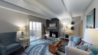 This thoughtfully designed single-floor space makes a strong first impression with its combination of architectural character and contemporary styling. The living area is the centerpiece of the home, showcasing exposed beam ceilings that add depth and visual interest overhead. Dark hardwood flooring runs throughout, grounding the space with warmth and contrast against the light grey walls.

A dramatic dark accent wall frames the fireplace, which serves as a natural focal point and creates a cozy, inviting atmosphere. A decorative mirror mounted above the mantle reflects light and enhances the sense of openness in the room. Ambient lighting from table lamps positioned throughout the space adds a soft, welcoming glow.

Natural light flows generously through a large sliding glass door, which leads to a private balcony — offering an outdoor retreat and views of the surrounding landscape. A tall indoor plant near the door brings a refreshing touch of greenery indoors.

The living space is finished with curated décor, including a plush blue and grey area rug that anchors the seating arrangement and introduces a pop of color. The overall ambiance is polished yet comfortable, blending modern design sensibilities with warm, livable details that make this a functional and visually appealing space.