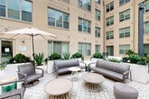 Located at 20013 Century Blvd in Germantown, Maryland, this thoughtfully designed outdoor amenity courtyard is set within a multi-story building featuring warm beige brick facades and expansive windows. The space is defined by its distinctive hexagonal paver flooring, which flows throughout the courtyard and creates a cohesive, modern aesthetic.

The courtyard is organized into several functional zones. A pergola-covered area adjacent to the pool provides shaded dining with light blue tables and chairs, offering a relaxing poolside atmosphere. The pool itself is visible through slatted privacy screens and a black iron fence, adding a resort-like quality to the setting.

A central open-air lounge area features contemporary wicker and upholstered sofas, swivel chairs, and stylish round coffee tables, all arranged to encourage social gathering. Large white planter boxes filled with agave, ferns, and tropical greenery provide natural definition between seating zones and add vibrant color to the space.

A dedicated grilling station with multiple barbecue units offers residents the convenience of outdoor cooking, complemented by additional shaded dining tables with umbrellas nearby. The multi-story building encloses the courtyard on multiple sides, creating a sheltered, semi-private atmosphere while allowing ample natural light to fill the space throughout the day.