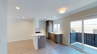 This single-floor space showcases a clean, modern design with an open-concept layout that seamlessly connects the kitchen and living areas. The kitchen is a standout feature, equipped with sleek two-tone cabinetry in white and warm wood-grain finishes, a stainless steel refrigerator, a dishwasher, and a gas cooktop set into a peninsula with a bright white countertop. The peninsula serves as both a functional cooking and prep space and a natural divider between the kitchen and the living area, encouraging an effortless flow throughout the unit. A dedicated sink area sits beneath a window, allowing natural light to brighten the workspace. Light-toned hardwood-style flooring runs throughout, adding warmth and continuity to the space. The ceilings are smooth and bright white, contributing to an airy, open atmosphere. Recessed lighting and a central flush-mount fixture provide well-distributed illumination. A sliding glass door opens directly onto a private balcony, offering outdoor access and allowing additional natural light to fill the interior. The overall ambiance is contemporary, bright, and functional, making it a practical yet stylish living space.