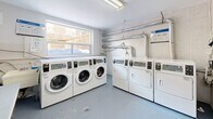 Located at 650 Woodbine Ave in Toronto's East End, this shared laundry facility is a compact, well-organized space designed for practical, everyday use. A narrow entry corridor with a drop-ceiling leads into the main laundry room, finished with durable grey flooring and clean white walls throughout.

The main area houses a bank of commercial-grade Huebsch front-loading washing machines, numbered for easy identification, positioned along one wall beneath a large window that brings in natural light and offers a view of the neighbouring outdoor area. Opposite, a set of matching dryers provides ample drying capacity. Exposed ventilation ductwork runs along the upper wall, underscoring the facility's functional design.

A folding table offers a convenient surface for sorting and organizing laundry. The space is equipped with a Sparkle digital payment kiosk, allowing residents to start machines via a mobile app or card, with instructional signage posted nearby. A water filtration unit and fire safety equipment are also present, reflecting attention to building standards.

The facility is bright, clean, and efficiently laid out, making it a straightforward and accessible amenity for residents of the building.