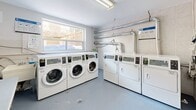 Located at 650 Woodbine Ave in Toronto's East End, this shared laundry facility is a compact, well-organized space designed for practical, everyday use. A narrow entry corridor with a drop-ceiling leads into the main laundry room, finished with durable grey flooring and clean white walls throughout.

The main area houses a bank of commercial-grade Huebsch front-loading washing machines, numbered for easy identification, positioned along one wall beneath a large window that brings in natural light and offers a view of the neighbouring outdoor area. Opposite, a set of matching dryers provides ample drying capacity. Exposed ventilation ductwork runs along the upper wall, underscoring the facility's functional design.

A folding table offers a convenient surface for sorting and organizing laundry. The space is equipped with a Sparkle digital payment kiosk, allowing residents to start machines via a mobile app or card, with instructional signage posted nearby. A water filtration unit and fire safety equipment are also present, reflecting attention to building standards.

The facility is bright, clean, and efficiently laid out, making it a straightforward and accessible amenity for residents of the building.