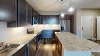 This single-floor unit in Wausau, Wisconsin, presents a well-designed layout with contemporary finishes and thoughtful details throughout. The kitchen serves as a standout feature, boasting rich dark-stained cabinetry extending to the ceiling, granite countertops, a central island with pendant lighting overhead, and a full suite of stainless steel appliances including a French door refrigerator, range, microwave, and dishwasher. Dark hardwood-style flooring and under-cabinet lighting complete the polished aesthetic.

The living area features the same hardwood-style flooring, a ceiling fan, and a sliding glass door that opens to a balcony, allowing natural light to flow in and providing outdoor access. Bedrooms are carpeted and painted in warm neutral tones, each equipped with ceiling fans. Corner placement and large, multi-panel windows in the primary bedroom offer expansive views of the surrounding Wausau landscape.

Bathrooms are finished with granite-topped dark vanities and wood-look flooring. One bathroom includes a tub/shower combination with a bright window, while another features a more private, corridor-style layout. A dedicated walk-in closet with wire shelving provides practical storage. Throughout the unit, clean white trim, neutral paint tones, and consistent finishes create a cohesive, modern ambiance.