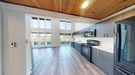 Located at 8500 20th Ave NE in Seattle's Ravenna neighborhood, this thoughtfully designed unit makes a striking impression with its dramatic double-height ceilings that amplify the sense of space well beyond its compact footprint. A standout architectural feature is the warm, natural cedar wood ceiling that crowns the open living area, adding texture and warmth to the otherwise clean, contemporary aesthetic.

Floor-to-ceiling windows along the exterior wall flood the main living space with abundant natural light and offer views of the surrounding neighborhood, while a sliding glass door opens to a private balcony. Light-toned hardwood-style flooring runs throughout, enhancing the airy feel of the space.

The kitchen is well-appointed with floor-to-ceiling dark wood-grain cabinetry, sleek white countertops, and a full suite of black appliances including a refrigerator, electric range, microwave, and dishwasher. A loft-style upper mezzanine, accessible via a compact staircase with metal railings, adds functional vertical space above.

The bathroom is clean and efficient, featuring a walk-in shower with white surround, a wall-mounted sink, and warm wood-look flooring. In-unit laundry is also present, adding everyday convenience. The overall ambiance is modern and bright, with industrial-inspired accents complementing the natural finishes throughout.