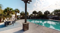 Located at 1735 Ashley Hall Rd in Charleston, South Carolina, this single-story property showcases a well-appointed outdoor amenity area designed for leisure and entertaining. The centerpiece is a generously sized swimming pool with clear turquoise water, framed by a spacious concrete deck ideal for sunbathing. Multiple lounge chairs are arranged along the poolside, offering ample space for relaxation beneath the open sky.

A standout feature of the outdoor area is a covered pergola structure supported by stone-base columns, providing a shaded retreat adjacent to the pool. Nearby, a built-in outdoor kitchen station features a stainless steel grill set into a stacked-stone base topped with polished dark granite countertop — a practical and attractive setup for outdoor dining and gatherings. Outdoor seating with modern rattan-style furniture and a patterned area rug completes the al fresco entertaining space.

The grounds are landscaped with tall Palmetto palms and manicured shrubs, reinforcing the distinctly Lowcountry, coastal ambiance characteristic of the Charleston area. Wrought iron fencing encloses the pool area, adding both security and a classic aesthetic. The overall outdoor environment conveys a resort-like atmosphere suited to the warm South Carolina climate.