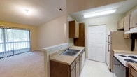 This single-floor residence in Tucker offers a functional and straightforward layout designed to maximize everyday comfort. The kitchen serves as a central feature of the home, showcasing warm-toned wood cabinetry with ample upper and lower storage. A double-basin stainless steel sink is set into a laminate countertop, complemented by a built-in dishwasher. The kitchen opens toward the living area via a half-wall peninsula, creating a natural flow between spaces and allowing for easy interaction between the kitchen and the adjacent room. Light-toned flooring in the kitchen transitions smoothly into carpeted living space, adding warmth underfoot. Overhead fluorescent lighting in the kitchen ensures a well-lit workspace. A set of bifold closet doors nearby provides additional storage, contributing to the unit's organizational practicality. The living area, visible through the open pass-through, features neutral carpeting and light walls, lending a clean and versatile backdrop. The textured ceiling is consistent throughout, adding subtle dimension to the interior. Overall, the space offers a comfortable and efficient single-level layout suitable for everyday living.