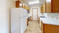 This single-floor unit offers a functional, well-organized layout with clearly defined living, sleeping, and kitchen zones. The living area features neutral wall-to-wall carpeting, warm wood baseboards, and a textured ceiling, creating a clean and adaptable space. A distinctive pass-through window with wood-trimmed cabinetry connects the living area to the kitchen, enhancing the sense of openness between the two spaces.

The galley-style kitchen is equipped with oak cabinetry extending to the ceiling for ample storage, white appliances including a full-size refrigerator and electric range, an over-range microwave, and a stainless steel sink. The flooring transitions to durable tile-style vinyl, and a door at the end of the kitchen provides additional access.

Multiple carpeted bedrooms each feature neutral tones, wood-trimmed windows with blinds that allow natural light, and textured ceilings with flush-mount lighting. The rooms offer generous wall space and are versatile in their potential use.

The bathroom includes an oak vanity with a wide white countertop, a large wall mirror, a toilet, and a walk-in shower with sliding glass doors. Overall, the unit maintains a consistent, neutral aesthetic throughout with warm wood accents providing cohesion.