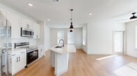 This well-appointed single-story home offers three bedrooms, two bathrooms, and a thoughtfully designed open-concept layout. The kitchen is a standout feature, boasting white shaker cabinetry with matte black hardware, a white subway tile backsplash, stainless steel appliances including a French door refrigerator, electric range, and built-in microwave. A central island with a quartz countertop, undermount sink, and striking black pendant lights serves as the focal point, flowing seamlessly into the bright living area, which is lined with windows that invite abundant natural light. Warm wood-look flooring extends throughout the main living spaces and hallway, complemented by crisp white walls and recessed lighting. The bedrooms feature plush carpeting and ceiling fans, with the primary bedroom offering generous proportions and ample natural light through wide windows. Three closets provide substantial storage, including a walk-in closet with built-in shelving and hanging rods. The two bathrooms feature white cabinetry, quartz countertops, and matte black fixtures. A dedicated laundry room houses a full-size washer and dryer. The long, light-filled hallway connects all rooms, reinforcing the clean, modern aesthetic that defines this move-in-ready home.