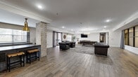Located at 120 Chubb Ave in Lyndhurst, NJ, this expansive single-floor amenity space offers a generously sized, open-concept layout that blends modern sophistication with industrial-chic design. Wide-plank hardwood-style flooring runs throughout, anchoring the warm, inviting atmosphere created by neutral walls, crown molding, and recessed lighting.

The space is thoughtfully divided into several distinct zones. A lounge area features tufted leather armchairs and a sofa arranged around a wall-mounted flat-screen TV, ideal for relaxed gatherings. Adjacent to it, a game area houses a full-size billiards table beneath an elegant chandelier, adding a recreational focal point. A dedicated dining zone includes a rustic wooden table with metal chairs, complemented by a modern linear pendant light and a wood-accented fireplace set into a stylish accent wall.

A full kitchenette runs along one side of the space, outfitted with sleek cabinetry, dark countertops, and brass pendant lighting, flanked by an exposed brick column that adds a striking architectural detail. Multiple windows throughout allow natural light to pour in, while views of the surrounding area enhance the open feel. The entry area provides a welcoming transition with clean lines and generous ceiling height, completing this well-appointed communal space.