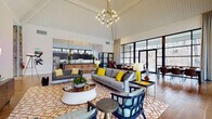 Located at 5146 N High St in Columbus, Ohio, this single-floor community space combines mid-century modern design with warm, functional interiors. The centerpiece of the space is a dramatic vaulted ceiling with exposed white-painted beams, anchored by a striking multi-bulb geometric chandelier that floods the room with warm light. A sweeping wall of floor-to-ceiling black-framed windows and glass doors lines one side of the space, offering abundant natural light and views of the surrounding wooded landscape. The main lounge area features a generous seating arrangement with a tufted leather sofa, colorful accent chairs in teal and navy, and bold yellow throw pillows set atop a patterned area rug. A wall-mounted flat-screen TV and understated media console anchor one end of the lounge. Adjacent to the lounge, a fully equipped kitchen with dark cabinetry, a mosaic tile backsplash, stainless steel appliances, and a bar-height island provides a functional gathering point. A dramatic natural stone accent wall adds texture and visual depth throughout the space. The dining and recreation area features warm hardwood-style flooring, a live-edge dining table with leather chairs, a ping-pong table, and additional high-top seating with vibrant blue chairs. Direct access to an outdoor patio extends the entertaining space further.
