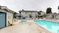 Located at 15134 Indiana Ave in Paramount, California, this residential complex offers a well-appointed communal outdoor amenity area as its centerpiece. The outdoor space is anchored by a generously sized swimming pool with clear blue water, complemented by an adjacent spa or hot tub — both enclosed within a metal safety fence. A spacious concrete pool deck surrounds the area, furnished with lounge chairs and Adirondack-style seating, providing ample space for relaxation and recreation.

The complex consists of single-story and two-story structures featuring classic horizontal lap siding in a neutral off-white palette, accented by blue-trimmed multi-pane windows and sliding glass doors that add a consistent, cohesive aesthetic throughout the property. Exterior staircases connect the upper and lower levels of the two-story buildings, and balconies offer additional private outdoor space for residents.

A dedicated clubhouse or common building is situated directly adjacent to the pool, featuring large multi-pane glass doors and decorative potted plants at the entrance. Mature trees and landscaping frame the perimeter of the complex, adding greenery and shade to the outdoor common areas. The overall ambiance is functional and community-oriented, reflecting a well-established California residential setting.