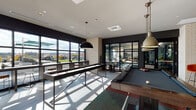 This contemporary amenity space in Watertown, MA is thoughtfully designed to offer a variety of social and recreational zones within a single, well-appointed floor. The layout flows seamlessly from one area to the next, unified by warm hardwood flooring, large-format tile, and a sophisticated dark-toned design palette.

The game room is a standout feature, equipped with a pool table, shuffleboard table, and adjacent bar area. Oversized windows and glass doors flood the space with natural light and provide direct access to an outdoor rooftop terrace, where scenic views of the surrounding Watertown landscape can be enjoyed.

Multiple distinct lounge areas cater to different needs. One seating zone features a large sectional sofa, pendant lighting with exposed Edison bulbs, and a backdrop of bold artwork visible through steel-framed glass partitions. A second lounge offers rich leather sectional sofas, a geometric accent wall in deep navy tones, and patterned area rugs that add warmth and texture. A third intimate nook is appointed with olive green armchairs, a round wood coffee table, curated wall art, and a built-in bookshelf with accent lighting.

A dedicated media and gaming room includes a round poker-style table, upholstered chairs, and multiple wall-mounted televisions. Exposed concrete columns and black steel-framed glass walls throughout lend the space an urban, loft-inspired character.
