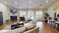 Located on Sweetwater Drive in Dothan, Alabama, this single-story community space is designed to provide a comfortable and inviting gathering environment. The main living area immediately impresses with soaring vaulted ceilings supported by decorative columns, creating an open and airy atmosphere. Warm-toned walls complement rich hardwood-style flooring that flows throughout the space, lending a cohesive and polished aesthetic.

The lounge area is anchored by a classic fireplace framed by a white mantel, with a wall-mounted television positioned above. Generous seating arrangements, including plush sofas and upholstered accent chairs, are arranged to encourage conversation and relaxation. French doors along one wall provide direct views of an outdoor pool area, allowing natural light to fill the interior.

A separate dining nook features a round table and dark wood chairs, offering a more intimate setting within the open layout. The kitchen is equipped with warm wood cabinetry, granite countertops, stainless steel appliances, and a breakfast bar with barstool seating—providing a fully functional space for events or everyday use. Recessed lighting throughout ensures consistent, comfortable illumination across all areas.