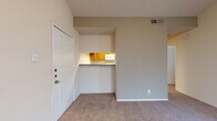 This single-story unit is defined by its vaulted ceilings, which lend a sense of openness and volume to the main living area. The living space is carpeted throughout and receives natural light through a glazed exterior door fitted with blinds, contributing to a bright and airy atmosphere. A second exterior entry door is positioned near a pass-through opening that connects the living area to the kitchen, providing both convenience and an open sightline between the two spaces.

The kitchen is equipped with warm oak cabinetry extending along two walls, offering ample storage. White appliances include a coil-top electric range, an over-the-range microwave, and a refrigerator. Countertops feature a speckled grey laminate surface, and the flooring transitions to a tile-pattern vinyl.

The bedroom is a well-proportioned room with soft carpet, a ceiling fan, and a large window that allows generous natural light to filter in while maintaining privacy with blinds.

The bathroom includes an oak vanity with a grey speckled countertop, an oval undermount sink, a wide wall mirror, and a full tub-shower combination with white tile surround. The textured ceilings and neutral wall tones carry consistently throughout the unit, creating a cohesive and functional living environment.
