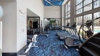 Located at 15250 Siesta Key Way in Rockville, Maryland, this fitness center is a well-appointed, single-floor facility designed to support a comprehensive workout experience. The space is immediately striking for its soaring double-height ceilings, accented by a bold blue ceiling detail and ceiling fans that keep the environment comfortable during intense sessions.

Floor-to-ceiling windows line one side of the facility, flooding the interior with natural light and offering open views of the surrounding community. The distinctive blue patterned carpet throughout adds a cohesive, modern aesthetic that ties the space together.

The main workout area is generously equipped with a full lineup of cardio machines, including treadmills, ellipticals, stationary bikes, and a rowing machine. A dedicated strength training zone features adjustable benches, a multi-station cable machine, a full dumbbell rack, and a medicine ball station with a variety of weights. Large mirrors are strategically positioned throughout, enhancing the sense of openness while allowing users to monitor their form.

A separate studio space, accessible through glass doors, is outfitted for group fitness or individual training, complete with stability balls and additional equipment. A dedicated storage unit near the entrance houses yoga mats, making the space fully ready for mat-based workouts. The overall ambiance is clean, energetic, and purpose-built for fitness.