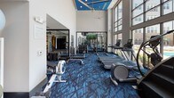 Located at 15250 Siesta Key Way in Rockville, Maryland, this fitness center is a well-appointed, single-floor facility designed to support a comprehensive workout experience. The space is immediately striking for its soaring double-height ceilings, accented by a bold blue ceiling detail and ceiling fans that keep the environment comfortable during intense sessions.

Floor-to-ceiling windows line one side of the facility, flooding the interior with natural light and offering open views of the surrounding community. The distinctive blue patterned carpet throughout adds a cohesive, modern aesthetic that ties the space together.

The main workout area is generously equipped with a full lineup of cardio machines, including treadmills, ellipticals, stationary bikes, and a rowing machine. A dedicated strength training zone features adjustable benches, a multi-station cable machine, a full dumbbell rack, and a medicine ball station with a variety of weights. Large mirrors are strategically positioned throughout, enhancing the sense of openness while allowing users to monitor their form.

A separate studio space, accessible through glass doors, is outfitted for group fitness or individual training, complete with stability balls and additional equipment. A dedicated storage unit near the entrance houses yoga mats, making the space fully ready for mat-based workouts. The overall ambiance is clean, energetic, and purpose-built for fitness.