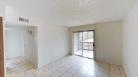 This single-floor space presents a straightforward and functional layout with a clean, neutral aesthetic throughout. The main living area is finished with light-toned ceramic tile flooring that extends across the space, contributing to a bright and airy feel. Walls are painted in a crisp white, enhancing the sense of openness and allowing natural light to reflect freely throughout the room. The ceiling features a classic textured popcorn finish, adding a familiar residential character to the interior. A prominent sliding glass door, dressed with vertical blinds, serves as both a focal point and a natural light source, providing direct access to an outdoor area framed by a wooden fence and mature greenery. This connection to the outdoors allows for views of the exterior surroundings and brings additional daylight into the living space. The overall ambiance is clean, minimal, and move-in ready, with a neutral palette that offers flexibility for a variety of furnishing and décor styles. The space is well-suited for comfortable everyday living, with its open feel and practical design elements making it an inviting and versatile environment.