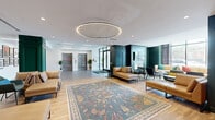 Located at 780 S 52nd St in the West Philadelphia neighborhood, this expansive lobby and communal lounge space offers a refined, hotel-inspired atmosphere across a generous single-floor footprint. The entrance foyer greets residents with large-format gray tile flooring, twin stainless steel elevators, floor-to-ceiling glass entry doors, and crisp white ceilings accented by linear lighting — creating a bright, welcoming arrival experience.

The space transitions seamlessly into a richly appointed lounge area anchored by warm hardwood flooring and a striking oversized circular LED pendant light. Deep teal and forest green wainscoting and paneled columns lend the space a bold yet sophisticated character, while tan leather sectionals and chaises arranged around an ornate area rug invite relaxed gathering.

A standout feature is the fully paneled green accent wall showcasing a decorative fireplace surround topped by a mounted flat-screen television — a elegant focal point that reinforces the upscale residential aesthetic. Adjacent seating nooks provide intimate lounge areas, including a windowed lounge with floor-to-ceiling views of the surrounding streetscape, navy accent walls, and eclectic seating arrangements. Throughout, curated artwork, indoor plants, and layered lighting complete the polished, community-oriented ambiance.