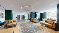 Located at 780 S 52nd St in the West Philadelphia neighborhood, this expansive lobby and communal lounge space offers a refined, hotel-inspired atmosphere across a generous single-floor footprint. The entrance foyer greets residents with large-format gray tile flooring, twin stainless steel elevators, floor-to-ceiling glass entry doors, and crisp white ceilings accented by linear lighting — creating a bright, welcoming arrival experience.

The space transitions seamlessly into a richly appointed lounge area anchored by warm hardwood flooring and a striking oversized circular LED pendant light. Deep teal and forest green wainscoting and paneled columns lend the space a bold yet sophisticated character, while tan leather sectionals and chaises arranged around an ornate area rug invite relaxed gathering.

A standout feature is the fully paneled green accent wall showcasing a decorative fireplace surround topped by a mounted flat-screen television — a elegant focal point that reinforces the upscale residential aesthetic. Adjacent seating nooks provide intimate lounge areas, including a windowed lounge with floor-to-ceiling views of the surrounding streetscape, navy accent walls, and eclectic seating arrangements. Throughout, curated artwork, indoor plants, and layered lighting complete the polished, community-oriented ambiance.