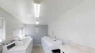 This single-floor laundry facility is designed with functionality and practicality at its core. The space is finished with clean white walls and a neutral gray accent wall at the rear, creating a bright and tidy environment. Multiple top-load washing machines are arranged in rows throughout the room, providing ample capacity for shared use. The machines are uniformly white, contributing to the space's organized and orderly appearance.

Overhead, large rectangular fluorescent light fixtures provide consistent, even illumination throughout the room, ensuring excellent visibility. The ceiling is smooth and white, lending a clean, no-frills aesthetic to the space. A countertop surface is visible along one side, offering a convenient area for sorting or folding laundry. A utility sink is also present, adding to the room's practical functionality.

The flooring appears to be a durable concrete or hard surface, well-suited for a high-traffic utility space. Informational and safety signage is posted on the walls, including a no-smoking notice. A waste bin is positioned near the entrance for convenience. Overall, the space is clean, well-lit, and efficiently organized for communal laundry use.