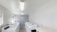 This single-floor laundry facility is designed with functionality and practicality at its core. The space is finished with clean white walls and a neutral gray accent wall at the rear, creating a bright and tidy environment. Multiple top-load washing machines are arranged in rows throughout the room, providing ample capacity for shared use. The machines are uniformly white, contributing to the space's organized and orderly appearance.

Overhead, large rectangular fluorescent light fixtures provide consistent, even illumination throughout the room, ensuring excellent visibility. The ceiling is smooth and white, lending a clean, no-frills aesthetic to the space. A countertop surface is visible along one side, offering a convenient area for sorting or folding laundry. A utility sink is also present, adding to the room's practical functionality.

The flooring appears to be a durable concrete or hard surface, well-suited for a high-traffic utility space. Informational and safety signage is posted on the walls, including a no-smoking notice. A waste bin is positioned near the entrance for convenience. Overall, the space is clean, well-lit, and efficiently organized for communal laundry use.