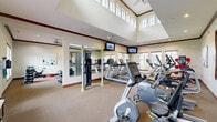 This well-appointed fitness center at 1021 Stoneridge Dr in Seymour, Indiana, is thoughtfully designed across two exercise rooms to support a comprehensive range of workout routines. The main exercise room is the architectural highlight of the space, boasting soaring double-height ceilings lined with a row of clerestory windows that flood the interior with natural light. Warm neutral tones, rich wood crown molding, and carpeted flooring create a polished yet comfortable environment. This primary room is equipped with Matrix cardio machines, including treadmills, ellipticals, and a stationary bike, alongside a HOIST cable machine for strength training. Wall-mounted flat-screen televisions and large mirrors further enhance the workout experience. A second, more intimate exercise room offers a dedicated space for functional and flexibility training, furnished with a spin bike, adjustable weight bench, dumbbells, medicine balls, stability balls, a trampoline, and a yoga mat. A wall-mounted television and a window with outdoor views make this room equally inviting. Together, the two rooms provide a versatile and fully equipped fitness facility suited for cardio, strength, and functional training needs.