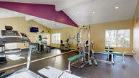 Located at 6152 Stanton Ave in Buena Park, California, this fitness center offers a well-equipped exercise space designed to support a range of workout routines. The room features a vaulted ceiling with a bold magenta accent that adds a vibrant visual element, while warm yellow walls and a durable rubberized floor contribute to a clean, functional atmosphere. Floor-to-ceiling mirrors line one wall, visually expanding the space and allowing users to monitor their form during exercise. Natural light filters in through multiple windows, with views of surrounding greenery and the outdoor pool area, creating a bright and inviting environment. The facility is equipped with a multi-station cable weight machine, a weight bench, a treadmill, a stationary bike, and a dumbbell rack, providing options for both strength training and cardiovascular workouts. A wall-mounted television is positioned for convenient viewing during exercise. Recessed lighting and a textured ceiling in the secondary section of the room ensure adequate illumination throughout. The space is thoughtfully laid out to maximize usability within its footprint, making it a practical and accessible fitness amenity for residents in Buena Park.