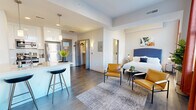 This 1-bedroom, 1-bathroom unit offers a thoughtfully designed open-concept layout that maximizes every square foot. The living and kitchen areas flow seamlessly together, anchored by warm hardwood-style flooring and bright, neutral walls throughout. Large windows dressed with bold red curtains invite generous natural light and offer views of the surrounding cityscape, enhancing the sense of openness.

The kitchen is a standout feature, equipped with full-height white cabinetry, a white tile backsplash, stainless steel appliances including a refrigerator, range, microwave, and dishwasher, and a peninsula with a white quartz countertop and bar seating. Pendant lighting above the peninsula adds a modern touch. The living area provides comfortable space for seating and entertainment.

The bedroom is a private, well-proportioned retreat featuring large windows with city views, a striking navy upholstered headboard, hardwood-style flooring, and warm ambient lighting from flanking bedside lamps. A closet with built-in hanging space is conveniently located nearby.

The bathroom is finished in crisp white marble-look tile, featuring a glass-enclosed walk-in shower, a stacked in-unit washer and dryer, built-in shelving niches, and ample towel storage — combining practicality with clean, modern aesthetics throughout.