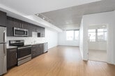This contemporary urban unit offers a well-designed single-floor layout that blends industrial and modern aesthetics throughout. The open-plan living and dining area flows seamlessly into the kitchen, creating a spacious, connected environment ideal for everyday living. Warm hardwood flooring spans the main living areas, contrasting beautifully with the exposed concrete ceiling — a standout architectural feature that lends the space an industrial character. Track lighting mounted along the ceiling provides adjustable illumination across the open area.

The kitchen is outfitted with dark espresso cabinetry extending to ceiling height, light-toned countertops, and a full suite of stainless steel appliances including a refrigerator, electric range, over-the-range microwave, and dishwasher. A dedicated in-unit laundry closet with a stacked washer and dryer adds everyday convenience.

Large windows in both the living area and bedroom draw in generous natural light while offering views of the surrounding urban cityscape. The bedroom features neutral carpet flooring and a clean, minimalist design. The bathroom is finished with a dark wood vanity, white countertop, a full tub-shower combination, and bright white tile surround. Overall, the space presents a polished, contemporary atmosphere suited to urban living.