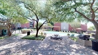 This outdoor amenity space centers around a generously sized swimming pool with clear turquoise water, framed by brick coping and a wide surrounding deck. The pool area is thoughtfully designed to accommodate both relaxation and social gathering, with multiple lounge chairs positioned along the poolside for sunbathing and leisure. A wooden pergola structure provides a shaded retreat near the water's edge, complemented by additional seating areas with tables and chairs suitable for outdoor dining or casual conversation. Mature trees throughout the space offer natural shade and a sense of established landscaping, enhancing the overall comfort of the environment. Neatly trimmed hedges and potted plants add greenery and visual appeal to the hardscaped surroundings. The backdrop consists of classic red brick multi-story residential buildings with pitched rooflines, lending the space a cohesive and well-maintained aesthetic. An open-sky setting ensures abundant natural light throughout the day, while the fenced perimeter provides privacy and security. Overall, this outdoor pool area presents a functional and inviting communal retreat designed for year-round enjoyment in a sunny climate.