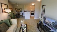 This single-floor residence offers an open and cohesive layout that seamlessly connects the main living areas. The living room serves as the heart of the space, featuring plush carpeting, a classic area rug, and a comfortable seating arrangement centered around a dark wood coffee table. Track lighting overhead provides versatile illumination, complementing the warm, inviting ambiance of the room. Adjacent to the living space, the dining area accommodates a full dining set and is accented by a large decorative mirror that adds depth and visual interest to the wall. The kitchen is positioned at the center of the floor plan, defined by a white peninsula counter that doubles as a functional workspace and casual divider between the cooking and living zones. White cabinetry extends to the upper walls, offering generous storage, while a white range and standard appliances complete the setup. Light-toned flooring in the kitchen contrasts subtly with the carpeted living and dining areas, helping to delineate each zone naturally. Neutral wall tones throughout create a clean, airy backdrop that enhances the sense of openness. Decorative touches, including floral arrangements and framed artwork, lend a polished, move-in-ready feel to this thoughtfully designed single-level home.