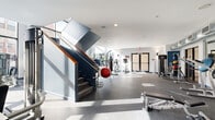 This fitness center is a thoughtfully designed, single-floor facility that offers a comprehensive workout environment suited for a wide range of exercise routines. The space is defined by its open, airy layout, with high ceilings and large floor-to-ceiling windows that flood the interior with abundant natural light and provide views of the surrounding outdoor area, including a pool courtyard.

The flooring throughout consists of durable speckled rubber, ideal for high-traffic fitness use. A prominent staircase with a navy blue frame and warm wood handrail serves as an architectural focal point, dividing the space into distinct workout zones while adding a modern, industrial aesthetic.

The strength training area is well-appointed with a full rack of dumbbells, adjustable benches, cable machines, and a variety of selectorized resistance equipment. A boxing station featuring a heavy bag and speed bag adds a dynamic training option. The cardio zone includes stationary bikes and additional cardio machines positioned to take advantage of the natural light from the expansive windows.

Wall-mounted mirrors amplify the sense of space and support form-checking during workouts. Exposed ductwork along the ceiling contributes to the facility's modern industrial character. Recessed lighting supplements natural light throughout. The space is accessible via glass-paneled double doors, maintaining a welcoming and open atmosphere throughout.