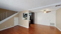 This two-story residence is designed with a practical, open layout that maximizes functionality across both levels. The main floor centers around a spacious living area finished with rich hardwood-style flooring and a ceiling fan overhead. A textured ceiling runs throughout the lower level, adding visual depth. The kitchen is accessible via a pass-through window and half-wall counter, creating a connected feel between the cooking and living spaces. Inside, the kitchen is outfitted with dark espresso cabinetry, light countertops, stainless steel appliances including a refrigerator, electric range, and over-the-range microwave, along with a utility area housing a stacked washer and dryer. A staircase along one wall leads to the upper level, where the bedrooms are carpeted and feature neutral warm-toned walls and vertical blind-covered windows that allow natural light to filter in. One bedroom includes sliding glass door access to an outdoor space. Two bathrooms are present, each featuring dark vanity cabinetry, light countertops, large mirrors, and coordinating brushed nickel fixtures. One bathroom includes a sliding glass shower enclosure.