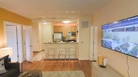 This well-appointed single-floor residence offers a thoughtfully designed open-concept layout that maximizes both space and functionality. The kitchen serves as a central focal point, featuring light maple cabinetry, dark countertops, and a full suite of stainless steel appliances, including a refrigerator, microwave, and range. A peninsula-style breakfast bar extends from the kitchen, lined with sleek chrome bar stools that create a casual dining and gathering area. Track lighting installed along the kitchen ceiling provides ample illumination, complementing the warm, inviting tones of the space. Rich hardwood flooring flows throughout the main living area, adding warmth and visual continuity. The open layout allows the kitchen to connect effortlessly with the living space, fostering a social and airy atmosphere. Neutral, warm wall tones throughout create a welcoming ambiance, while the clean, modern aesthetic is reinforced by white trim, panel doors, and contemporary furnishings. A wall-mounted flat-screen television is positioned in the living area, enhancing the space's modern appeal. The overall design balances practicality with style, making it well-suited for everyday living and entertaining alike.