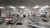 This dedicated fitness center is thoughtfully laid out to accommodate a full range of exercise routines. The main workout area features durable carpet flooring throughout, providing a comfortable and functional surface for various activities. Fluorescent lighting illuminates the space evenly from above, complemented by a textured ceiling that adds subtle visual interest.

The cardio section is well-stocked with multiple treadmills and elliptical trainers, each equipped with individual monitor screens for entertainment during workouts. A stationary bike rounds out the cardio offerings. For strength training, the facility includes a multi-station Life Fitness cable machine, a dedicated weight bench, and a full dumbbell rack with an organized selection of free weights.

Full-length mirrors line the walls, visually expanding the space and allowing users to monitor their form. A pair of analog wall clocks are mounted for easy time-tracking during sessions. The entry area transitions from tile flooring to the carpeted workout zone, with warm wood-toned doors leading to adjacent rooms, suggesting access to changing or restroom facilities nearby. The overall layout is practical and efficiently organized, making effective use of the available space to deliver a comprehensive fitness environment.