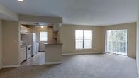 This single-floor residence offers an open-concept layout that combines the living and kitchen areas into one cohesive, functional space. The living area is generously sized, featuring neutral-toned carpet flooring and warm beige walls that create a calm, welcoming atmosphere. Large windows along the exterior wall flood the space with abundant natural light, and a sliding glass door provides direct access to a private outdoor balcony, offering views to the outside.

The kitchen is thoughtfully positioned at the heart of the unit, defined by a raised peninsula counter that provides a natural division between the cooking and living spaces while maintaining an open feel. The kitchen features upper cabinetry in a light finish, a full-size refrigerator, and additional appliances, all set against a tile floor that contrasts with the carpeted living area. An overhead light fixture illuminates the kitchen workspace, while a window above the sink brings in additional natural light.

The recessed soffit detail above the kitchen adds architectural interest and helps delineate the space. The overall design is clean, practical, and move-in ready, with a neutral palette that offers flexibility for a variety of interior styles. The abundant windows and open flow between areas give the residence a spacious, airy quality throughout.