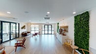 Located at 12950 SW 127th Avenue in Miami, FL, this amenity floor offers a thoughtfully designed collection of spaces built for comfort, community, and wellness. The lobby corridor makes an immediate impression with warm wood-look tile flooring and a striking cluster of oversized woven rattan pendant lights, leading residents toward bright, open gathering areas. The main lounge is an airy, light-filled space featuring large glass doors that open to an outdoor balcony with water views, modern seating arrangements with leather sofas and accent chairs, living green wall panels, hexagonal wall shelves, and a sleek refreshment station. The overall ambiance is fresh and welcoming, blending natural textures with clean, contemporary lines. A dedicated specialty room features built-in cabinetry with ample storage and dramatic textured wave-pattern wall panels in deep navy, creating an intimate and acoustically considered environment. The fitness center is generously sized and well-equipped with treadmills, a rowing machine, cable machines, a multi-station strength unit, and additional free-weight and functional training equipment. Large windows allow abundant natural light into the gym, which also features ceiling fans, rubberized flooring, and motivational wall graphics. Restrooms and a water fountain station serve the fitness area, completing this comprehensive amenity offering.